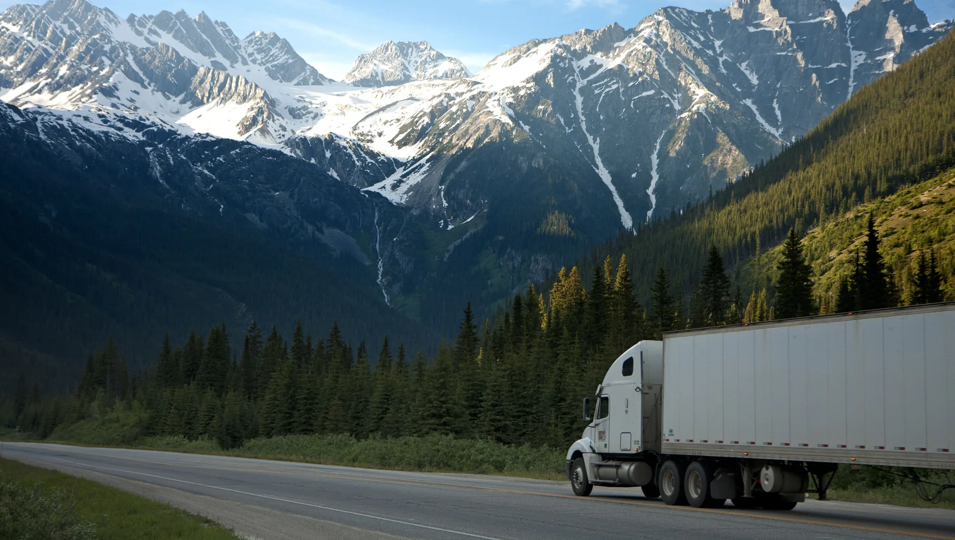 White semi-truck on a mountain highway beneath snow-capped peaks