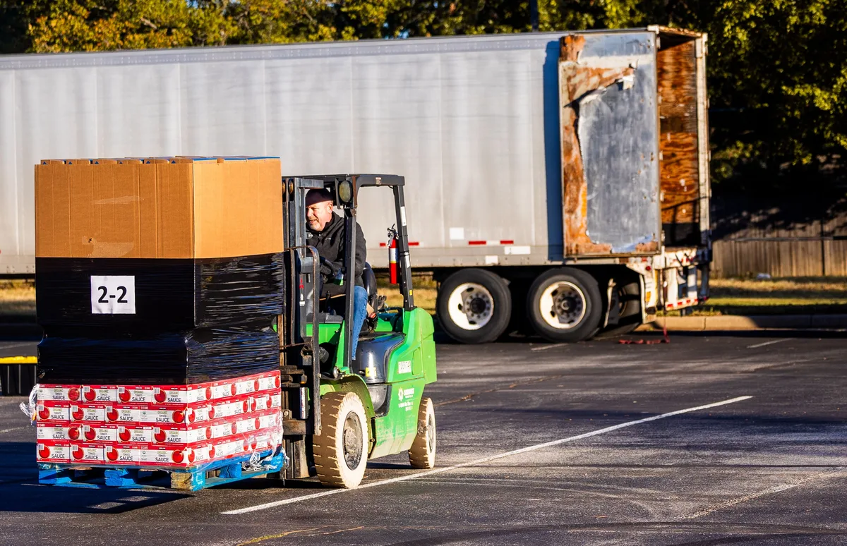 Forklift carrying a shrink-wrapped pallet of boxed goods beside an open trailer